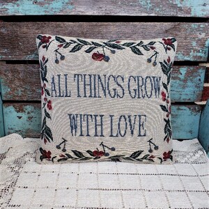 Puede incluir: Una almohada decorativa con fondo crema y un borde floral. La almohada presenta el texto "All things grow with love" en bordado azul.