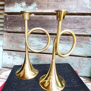 May include: Two polished brass candle holders shaped like miniature trumpets. Each holder features a wide, flared base, a slender vertical stem, a circular loop design, and a small cup at the top designed to securely hold a single taper candle.