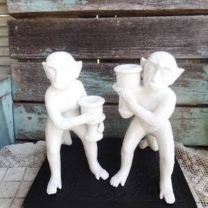 May include: A pair of white ceramic monkey candle holders. Each monkey is holding a candle holder in the shape of a cup. The candle holders are sitting on a black book.