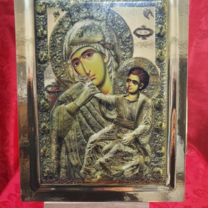 Può includere: Un'icona religiosa dorata che raffigura la Vergine Maria e il bambino Gesù. L'icona è incorniciata in una cornice dorata con dettagli decorativi. L'icona è su un cavalletto di legno.