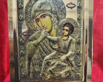 May include: A gold-toned religious icon depicting the Virgin Mary and baby Jesus. The icon is framed in a gold-colored frame with ornate details. The icon is on a wooden easel.