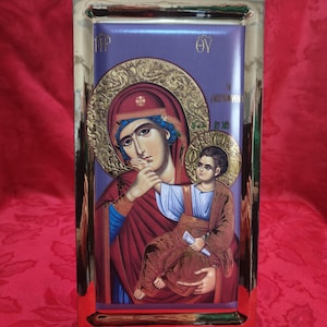 Può includere: Un'icona religiosa incorniciata che raffigura la Vergine Maria che tiene in braccio il bambino Gesù. L'icona è dipinta in uno stile tradizionale con accenti dorati e uno sfondo viola. Il testo "MP OY" è visibile negli angoli superiori della cornice.