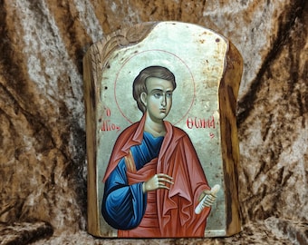 May include: A hand-painted icon of a young man with a halo, holding a scroll. The icon is painted on a wooden panel with gold leaf accents. The text "Ο Άγιος Θωμάς" is written in Greek on the icon.