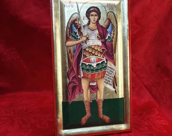 May include: A framed icon of an angel with wings, holding a sword and a scroll. The angel is depicted in a red and gold robe, with a green background. The icon is framed in gold and red.