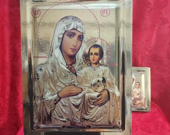 May include: A rectangular gold-toned metal icon plaque featuring a raised relief depiction of a mother and child. The figures are adorned in detailed robes with halos. Greek lettering is visible in the upper corners of the metallic surface.