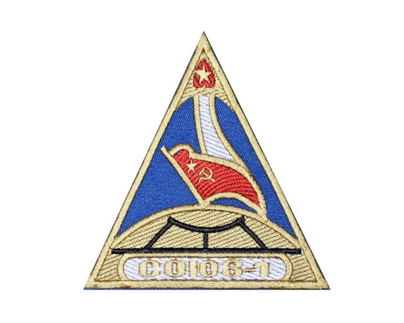 May include: Embroidered patch in a triangular shape with a blue background and gold trim. The design includes a red flag with a star and hammer and sickle, a bridge, and the text "CO106-1".