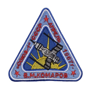Może przedstawiać: Niebieska, trójkątna naszywka z białym obramowaniem i czerwoną gwiazdą w każdym rogu. Naszywka przedstawia stylizowane zdjęcie statku kosmicznego z słońcem i promieniami w tle. Tekst "СОЮЗ СССР АПРЕЛЬ 1967 г. В.М. КОМАРОВ" jest wyszyty na czerwono i biało.