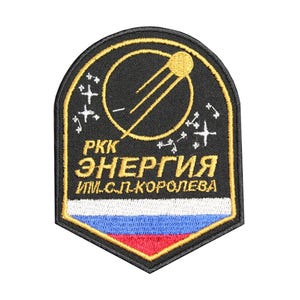 May include: A black and gold embroidered patch with the text "РКК ЭНЕРГИЯ им. С.П. КОРОЛЕВА" and a stylised image of a satellite orbiting a planet. The patch features the Russian flag in the bottom portion.