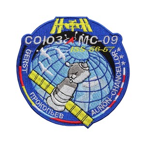 May include: A blue and yellow embroidered patch with the text "Soyuz MC-09 ISS 56-57" and the names "Gerst", "Prokopyev", and "Ayonin-Chancellor" around a globe with a space shuttle.