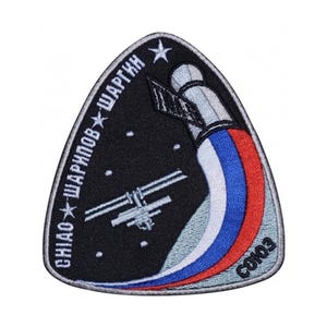 May include: A black and grey embroidered patch with a white star and the text "ГНИАО - ШАРПНОВ - ШАРГЫН" in Russian. The patch features a stylised image of a spacecraft docked with the International Space Station, with a red, white and blue ribbon trailing behind it. The text "СОЮЗ" is also visible on the patch.
