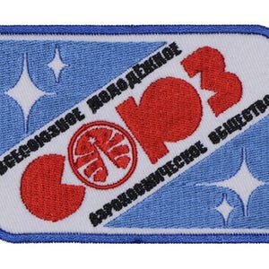 May include: Embroidered patch with a blue and white background, featuring red text and a circular emblem. The patch has a rectangular shape with rounded corners and a blue border. The text includes Cyrillic characters.