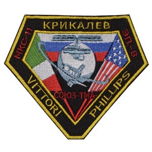 May include: A black and yellow embroidered patch with the text "KRIKALEV", "VITTORI", "PHILLIPS", "SOYUZ-TMA", "EP-8", and "MIKSI" in a diamond shape. The patch also features a space shuttle, the Russian and American flags, and the Italian flag.