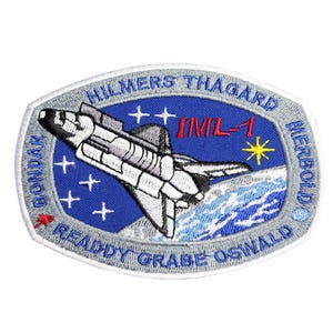 Könnte beinhalten: Ein weiß-blauer gestickter Aufnäher mit einem Raumschiff, Sternen und dem Text "IML-1" in der Mitte. Der Text "HILLMERS-THAGARD" befindet sich oben auf dem Aufnäher, und der Text "GONDAK READY GRABE OSWALD MERBOLD" befindet sich unten auf dem Aufnäher.