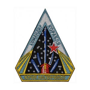 Op de afbeelding: Geborduurde patch in een driehoekige vorm met een ruimtethema. De patch toont een raket die de sterrenhemel in lanceert, een rode ster en tekst in het Russisch. De rand is lichtblauw en geel.