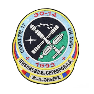 May include: A circular embroidered patch with a blue, white and red border. The centre of the patch is black with a white and grey space station design. The text "30-14", "СОЮЗ-ТМ-17", "ОК-МИР", "1993", "ЦИБЛИЕВ В.В. СЕРЕБРОДА", and "Ж.-П. ЭНЬЕРЕ" is embroidered in yellow, white and red.