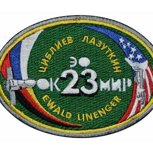 Op de afbeelding: Een groene, ovale patch met de tekst "Юбилей Лазуткин" en "Ewald Linenger" rond de rand. Het midden van de patch toont het nummer "23" en een gestileerde afbeelding van een ruimtestation.