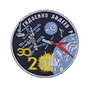 Puede incluir: Un parche circular bordado con fondo azul oscuro. El parche presenta una imagen blanca y azul de la Estación Espacial Internacional, un cohete rojo, un sol amarillo y una Tierra azul. El texto "ГІДЗЕНКО АВДЄЄВ РАНЬЄР" está escrito en un círculo blanco alrededor de la imagen. El número "302" está escrito en amarillo debajo de la imagen.