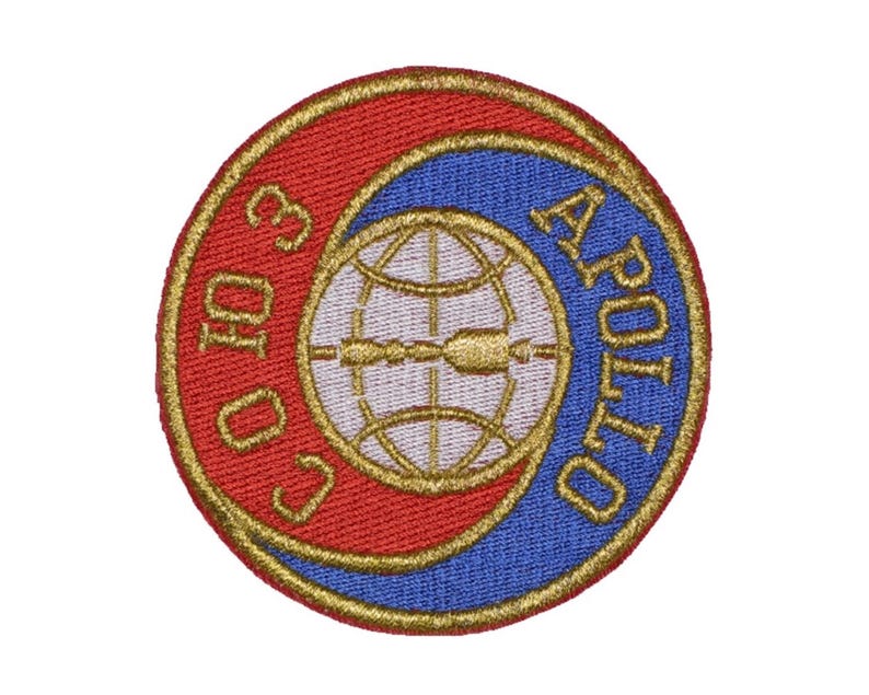 Pu&ograve; includere: Una toppa rotonda ricamata con sfondo rosso e blu. La toppa presenta un globo dorato con una freccia stilizzata e il testo "APOLLO 10" in oro.