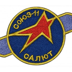 Puede incluir: Un parche bordado azul y dorado con una estrella roja y el texto "СОЮЗ-11 САЛЮТ" en escritura cirílica.