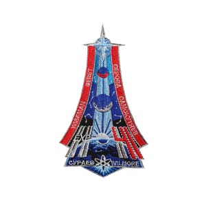 May include: A blue and silver space mission patch with red and white stripes. The patch features a rocket launching into space with the names of the astronauts: Wiseman, Gerst,  Samouylov,  Cerova,  Supaev, and Willmore.