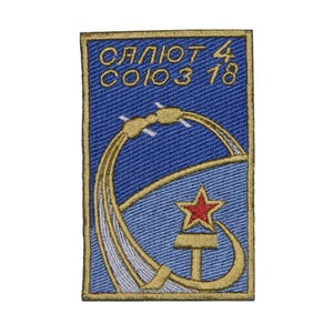 Puede incluir: Un parche bordado azul y dorado con el texto "СЛЁТ 4 СОЮЗ 18" y una estrella roja. El parche representa un cohete que se lanza al espacio.