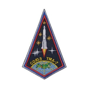 Puede incluir: Un parche triangular con un borde azul y un fondo azul oscuro. Un cohete blanco con una llama amarilla se encuentra en el centro del parche. El texto "СОЮЗ ТМА-7" está en la parte inferior del parche.