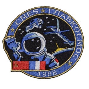 Könnte beinhalten: Ein blauer und goldener gestickter Aufnäher mit einer Weltraumszene. Der Aufnäher zeigt einen Weltraumspaziergang mit einem Astronauten, einer Raumstation und Sternen. Der Text "СНЕС ГЛАВКОСМОС" ist in einem Kreis um die Szene geschrieben. Das Jahr "1988" ist unten auf dem Aufnäher geschrieben.