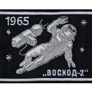 Op de afbeelding: Een geborduurde patch in zwart en wit met een witte astronaut in een ruimtepak dat in de ruimte zweeft. De patch heeft de tekst "1965" en "ВОСХОД-2" in wit.