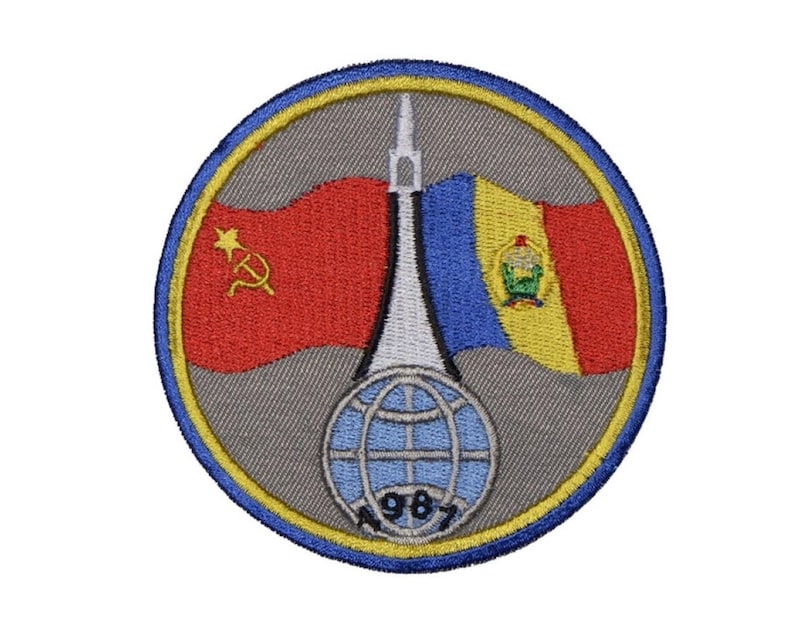 May include: A circular embroidered patch with a gray background and a yellow border. The patch features a white rocket, a red and yellow flag with a hammer and sickle, a blue and yellow flag with a coat of arms, and a globe with the year 1987.