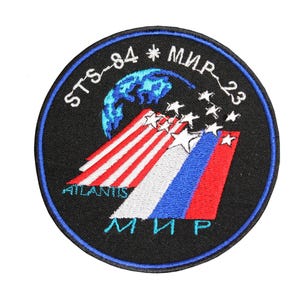 以下が含まれることがあります： 黒と青の刺繍パッチで、テキスト「STS-84 * MIR-23」と「ATLANTIS」が白で書かれています。パッチには、赤、白、青のストライプ模様と星がついた青と白の地球儀が描かれています。