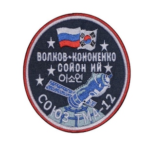 Puede incluir: Parche bordado redondo con fondo azul marino y borde rojo y blanco. Presenta una ilustración de nave espacial, las banderas de Rusia y Corea del Sur, y texto en cirílico y coreano. Parche conmemorativo.