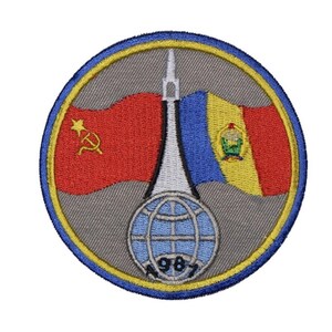 May include: A circular embroidered patch with a gray background and a yellow border. The patch features a white rocket, a red and yellow flag with a hammer and sickle, a blue and yellow flag with a coat of arms, and a globe with the year 1987.