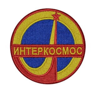 Puede incluir: Un parche bordado con un cohete estilizado lanzándose al espacio, con un esquema de colores rojo, amarillo y azul. El texto "ИНТЕРКОСМОС" está escrito en letras amarillas en el centro del parche.