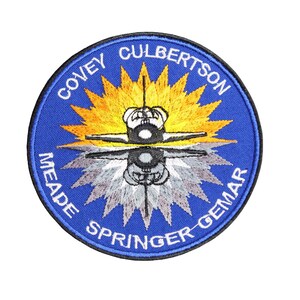 Puede incluir: Un parche bordado azul y blanco con los nombres "Covey Culbertson Meade Springer-Gemar" alrededor del borde. Una imagen estilizada de una lanzadera espacial está en el centro del parche.