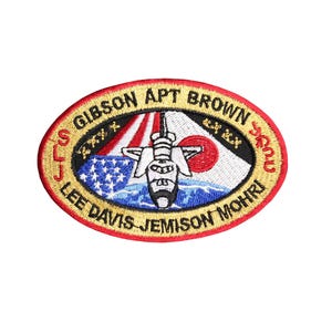 May include: A gold and red oval embroidered patch with the text "STS-47" and "SLJ" on the left side, "GIBSON APT BROWN" on the top, and "LEE DAVIS JEMISON MOHRI" on the bottom. The patch features a space shuttle in flight with the American and Japanese flags in the background.