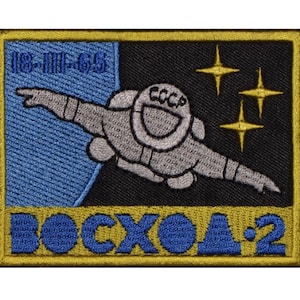 May include: A gold embroidered patch with a blue background and a black and white illustration of a cosmonaut in space. The patch reads "18-III-65" and "ВОСХОД-2" in Cyrillic.