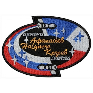 May include: A black and white embroidered patch with a blue, white and red background. The patch features a Soyuz spacecraft with the text "СОЮЗ-ТМ33 Афанасьев  Haluzhere  Казеев  СОЮЗ-ТМ32" in gold lettering.