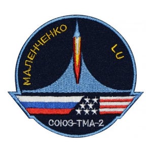 May include: A blue and white embroidered patch with a rocket ship design. The patch has the text "МАЛЫШЕНКО" and "LU" on the top and "СОЮЗ-ТМА-2" on the bottom. The patch also features the Russian and American flags.