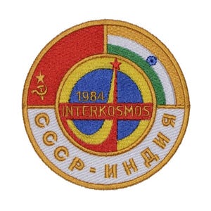 Peut inclure: Un écusson rond brodé avec un fond blanc et une bordure dorée. Au centre se trouve un globe bleu avec une croix rouge, jaune et verte. Le texte "INTERKOSMOS" est écrit en or au-dessus du globe, et "1984" est écrit en or en dessous du globe. Le texte "CCCP - ИНДИЯ" est écrit en or autour du globe. L'écusson est un symbole du programme spatial soviéto-indien.