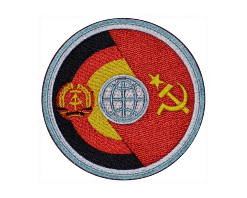 May include: A round embroidered patch with a red, yellow, and black design. The patch features a globe with a hammer and sickle on the right side and a symbol with a wreath on the left side.