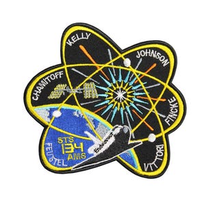 Op de afbeelding: Een geborduurde zwarte en gele patch met een spaceshuttle, een gestileerd atoom en de namen van de astronauten: Kelly, Johnson, Chamitoff, Fincke, Feustel en Vittorio. De tekst "STS-134" is ook zichtbaar op de patch.