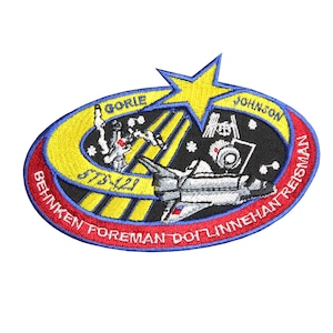 Puede incluir: Un parche bordado en blanco y negro con una estrella amarilla y una lanzadera espacial. El texto en el parche dice "STS-128" y los nombres de los astronautas: "Behnken, Foreman, Doty, Linnehan, Reisman, Gorrie, Johnson".