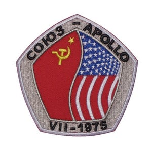 Peut inclure: Un &eacute;cusson brod&eacute; argent&eacute; et gris avec le texte "Soyouz - Apollo VII-1975" et un drapeau rouge et blanc avec une faucille et un marteau et des &eacute;toiles.