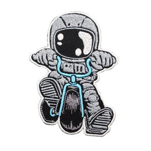 Op de afbeelding: Gestikte patch van een cartoon-astronaut in een grijs ruimtepak dat een blauwe driewieler rijdt. De astronaut heeft een zwart vizier en een witte helm.