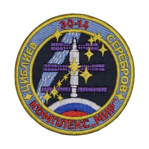 May include: A round, blue and gold embroidered patch with the text "30-14", "ЦИБЛЮЕВ", "СЕРЕБРОВ", and "КОМПЛЕКС "МИР" in red and yellow lettering. The patch features a stylised image of the Mir space station with stars and a blue and white background.