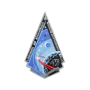 Puede incluir: Un parche triangular azul y blanco con un cohete lanzándose al espacio, una luna creciente y estrellas. El texto "LINDGREN 2018 KONONENKO VOLKOV-KORNIENKO KELLY 45" está impreso alrededor del borde del parche.