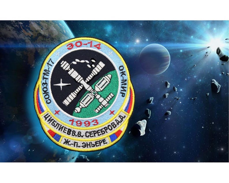 May include: A circular embroidered patch with a black background and a white outline. The patch features a space station with a green and white color scheme. The text "СОЮЗ-ТМ-17 30-14 ОК-МИР 1993 ЦИБЛИЕВ В.В. СЕРЕБРОВА А.А. Ж.-П. ЭНЬЕРЕ" is embroidered in white and yellow around the space station.