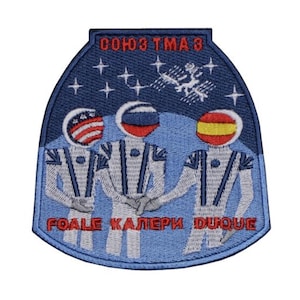 Puede incluir: Un parche bordado azul y blanco con el texto "СОЮЗ ТМА-3" y "ГОАЛЕ КАЛЕРИ ДУКУЕ" en rojo. El parche presenta tres astronautas con trajes espaciales con banderas en sus cascos: estadounidense, ruso y español. Los astronautas están de pie frente a un fondo azul con estrellas y una estación espacial.