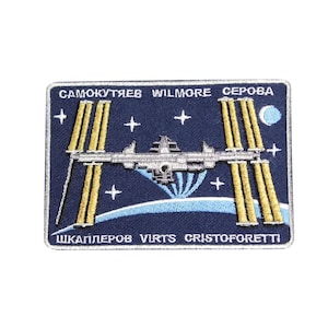 Puede incluir: Un parche bordado azul con los nombres de los miembros de la tripulación de la Estación Espacial Internacional: Samokutyaev, Wilmore, Serova, Shkaplerov, Virts y Cristoforetti. El parche presenta una estación espacial blanca orbitando una Tierra azul y blanca.