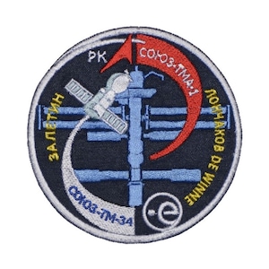 May include: A circular embroidered patch featuring a blue and white image of a space station with a red arrow pointing to the right. The text "СОЮЗ-TMA-1 ЛОНЧАКОВ DE WINNE" is written in a circle around the image. The text "ЗАЛЕТИН" is written on the left side of the image. The text "PK" is written at the top of the image. The text "СОЮЗ-TM-34" is written at the bottom of the image.